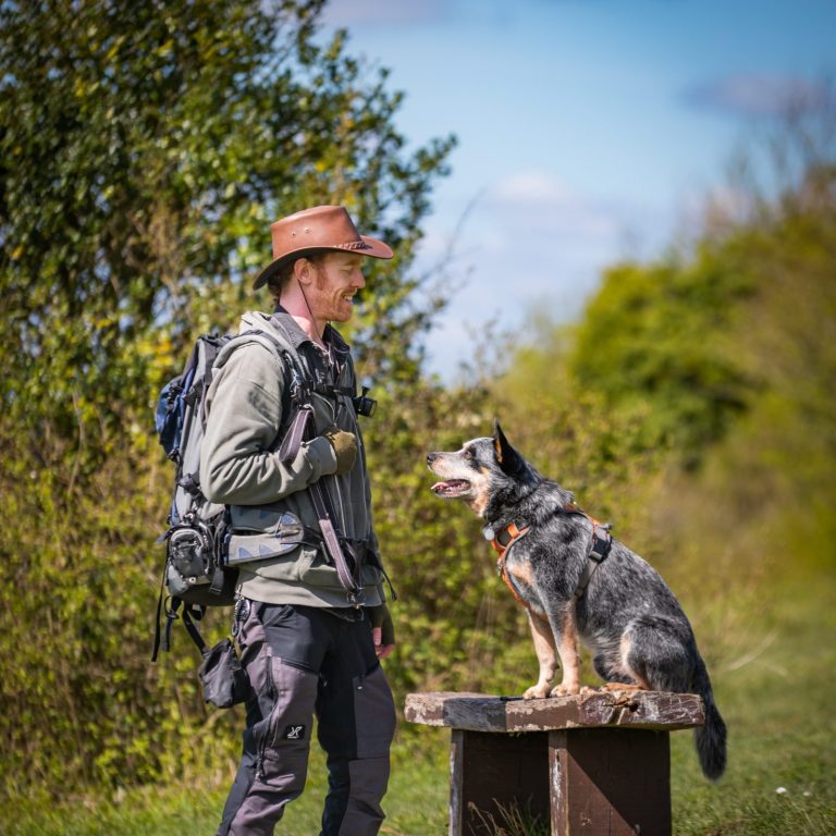 me and neo A person in a hat stands next to a dog on a bench in a lush outdoor setting.